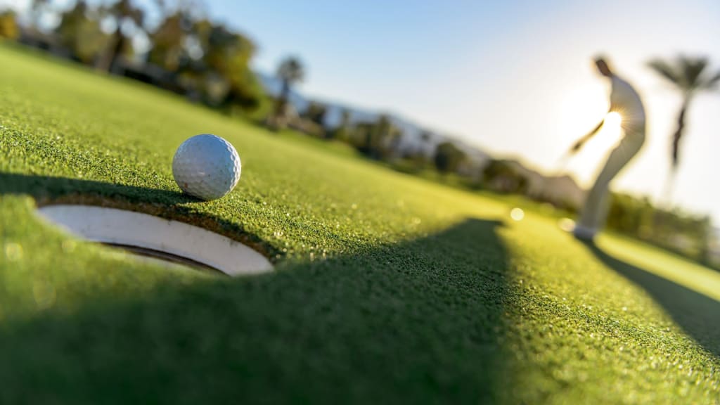 A close-up of a golf ball near the hole on a green, with a golfer in the background preparing to putt under a clear sky.